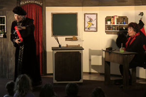 Der Zauberlehrer steht im Klassenraum der Hexenschule, mit dem Zauberbuch in der rechten Hand. Er spricht mit den Kindern im Publikum. Hexe Schmusemund sitzt erwartungsvoll an ihrem Pult.