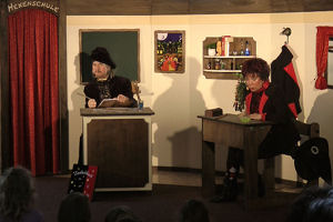 Zauberlehrer und Hexe Schmusemund sitzen an ihren Pulten und sprechen mit den Kindern im Publikum.