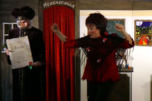 Der Zauberlehrer verleiht Hexe Schmusemund die Urkunde zur bestandenen Hexenprüfung. Die Hexe tanzt vor Freude.