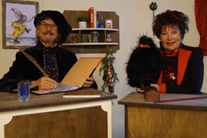 Der Zauberlehrer und Hexe Schmusemund an ihren Pulten in der Zauberschule. Der Zauberlehrer schreibt ins Zauberbuch. Hexe Schmusemund hat den Raben Flux auf dem Tisch sitzen.
