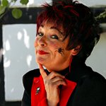 Hexe Schmusemund (Karin Meier) lächelt – mit roten Lippen, dichten schwarz-roten Haaren und einer schwarzen Spinne auf der linken Wange.