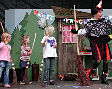 Hexe Schmusemund feiert eine bunte Geburtstagsparty vor ihrem Hexenhäuschen im Wald. Sie tanzt mit drei Kindern auf der Bühne – jeder mit einem Hexenreisigbesen in der Hand.